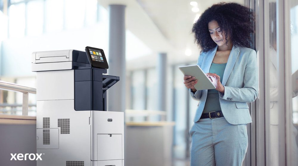 Xerox® ConnectKey®: Mobile Konnektivität Eine Person steht neben einem Xerox VersaLink B605-Multifunktionsdrucker, betrachtet ein Tablet-Gerät und interagiert damit. Dabei wird veranschaulicht, wie ConnectKey die mobile Interaktion mit dem Drucker ermöglicht.