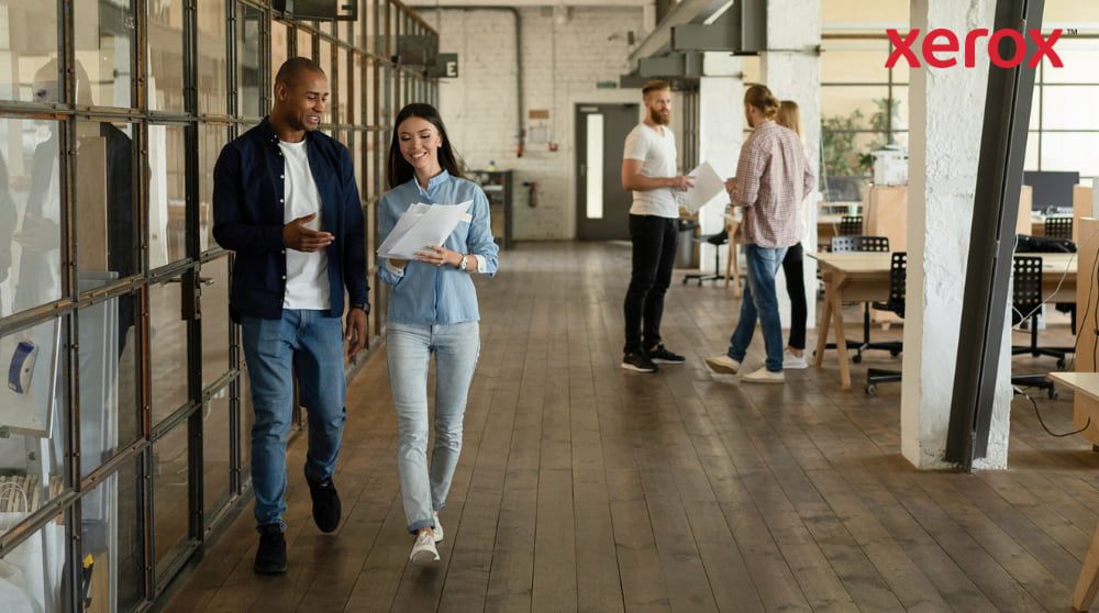 Zwei Kollegen gehen lächelnd einen Flur in einem modernen Großraumbüro entlang, halten Dokumente in den Händen und sind im Hintergrund mit weiteren Kollegen zu sehen, was einen dynamischen und kollaborativen Arbeitsplatz darstellt.