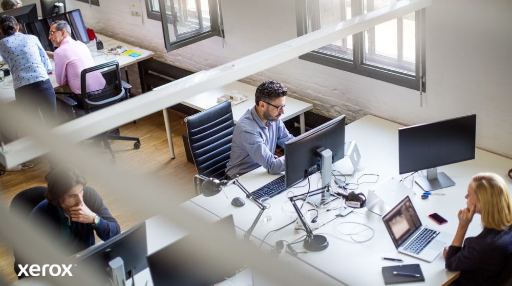 Xerox® Druck- und Scan-Erlebnis: Steigerung der Büroproduktivität Eine Draufsicht auf einen Großraumbüroraum mit mehreren Personen, die an ihren Schreibtischen am Computer arbeiten. Die Szene vermittelt den Eindruck einer produktiven und kollaborativen Arbeitsumgebung.