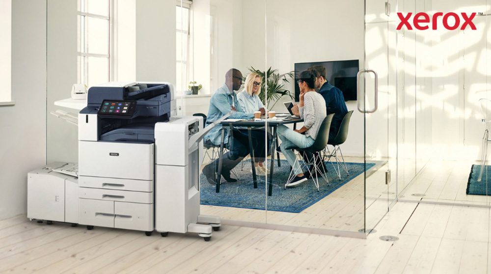 Im Vordergrund eines hellen Büros steht ein moderner Multifunktionsdrucker Xerox AltaLink B8245. Im Hintergrund sitzt eine Gruppe von Menschen an einem Tisch und diskutiert gemeinsam.