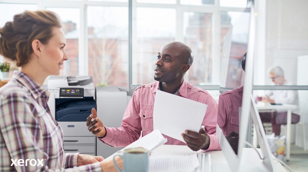 Eine Person überprüft Daten auf einem Laptop-Bildschirm mit einer digitalen Überlagerung verschiedener Gerätesymbole (Drucker, Mobilgeräte) und verbundener Linien, die das zentralisierte Flottenmanagement für Xerox Workplace Solutions symbolisieren.